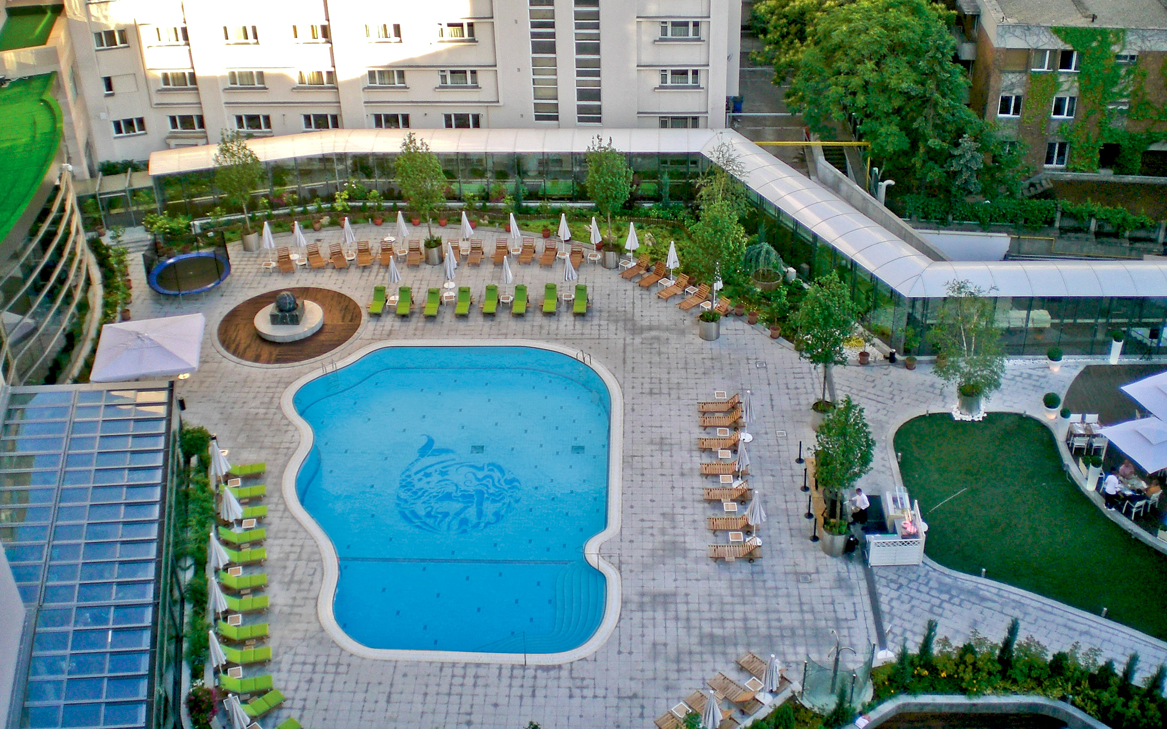The heated outdoor swimming pool is located on the underground parking garage of the hotel. Bird's eye view onto a courtyard with sun beds and a swimming pool